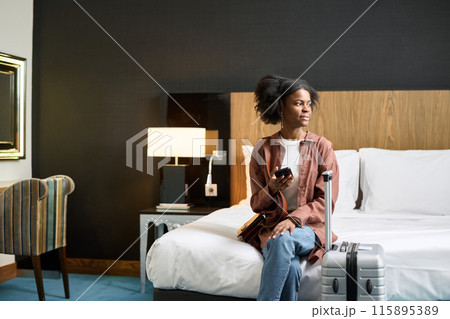 Portrait of young African American woman sitting on bed in hotel room and looking at window upon arrival copy space Portrait of young African American woman sitting on bed in hotel room and looking at window upon arrival copy space 115895389