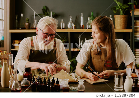 Senior perfumer sorting dried daisies and laughing while his female intern smiling widely and writing down perfumes recipe Senior perfumer sorting dried daisies and laughing while his female intern smiling widely and writing down perfumes recipe 115897804