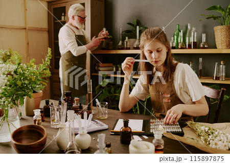Female perfumer sitting at table and smelling tester while mature male perfumer reading notes Female perfumer sitting at table and smelling tester while mature male perfumer reading notes 115897875