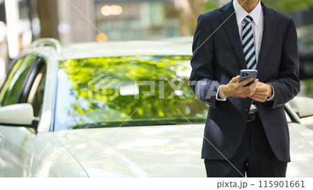 スマホを手に車の前に立つスーツの若い男性 115901661