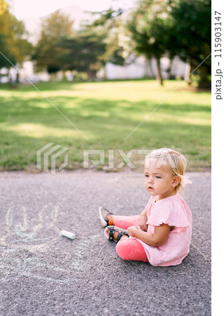 Little girl with chalk sits on the pavement in the park. High quality photo 115903147
