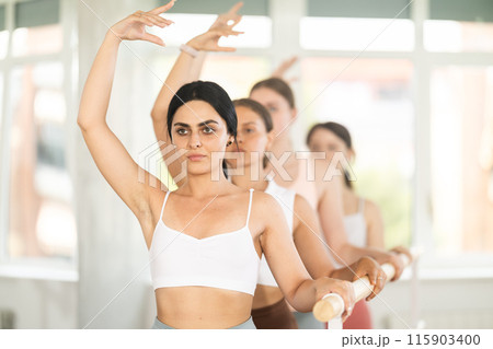 Portrait of pretty young girl practicing 3rd position of ballet stance in classical ballet with group of young beautiful girls in sports clothing in ballet hall 115903400