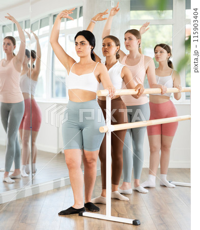 Experienced young female ballet dancer, dressed in sportswear and group of happy girls engaged in classical ballet performs exercise standing in 3rd position near ballet barre in dance hall Experienced young female ballet dancer, dressed in sportswear and group of happy girls engaged in classical ballet performs exercise standing in 3rd position near ballet barre in dance hall 115903404