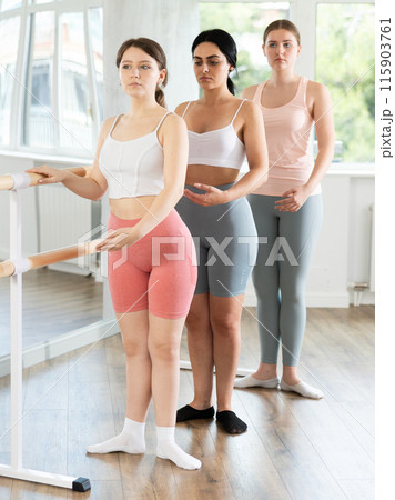 Happy young girl in sportswear, participating first position of ballet stance at barre with group youth people standing in row in choreography studio Happy young girl in sportswear, participating first position of ballet stance at barre with group youth people standing in row in choreography studio 115903761