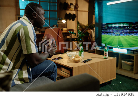 Young African American man holding ball sitting on couch in living room watching football game on TV in evening Young African American man holding ball sitting on couch in living room watching football game on TV in evening 115914939