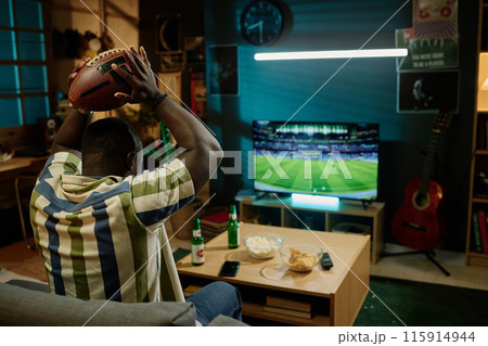 Unrecognizable young African American football fan holding ball above his head watching match on TV in living room, copy space 115914944