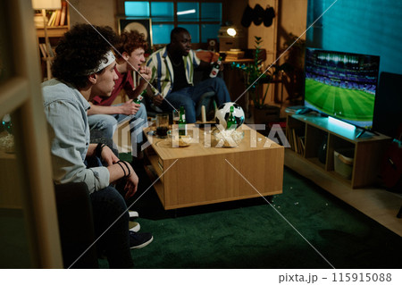 Group of multi-ethnic male friends sitting in living room watching soccer match on TV in evening, copy space Group of multi-ethnic male friends sitting in living room watching soccer match on TV in evening, copy space 115915088