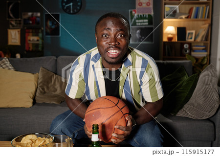 Medium shot of cheerful African American guy sitting on couch watching basketball match on TV late in evening Medium shot of cheerful African American guy sitting on couch watching basketball match on TV late in evening 115915177
