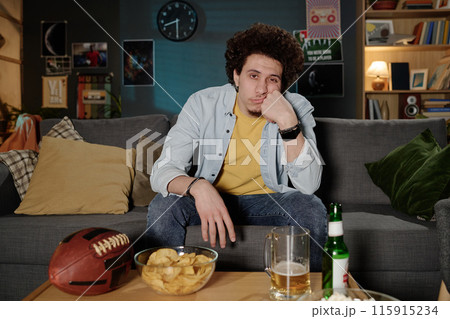 Sleepy young biracial man sitting on couch in living room watching football match on TV at night, copy space 115915234