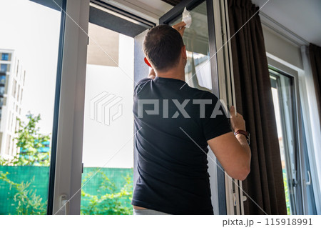 Young man cleans the windows in the house. He splashes detergent, rubs and removes water at the glass Young man cleans the windows in the house. He splashes detergent, rubs and removes water at the glass 115918991