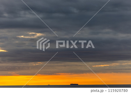 The sun is setting over the ocean with ships in the distance, under a dramatic sky The sun is setting over the ocean with ships in the distance, under a dramatic sky 115920780