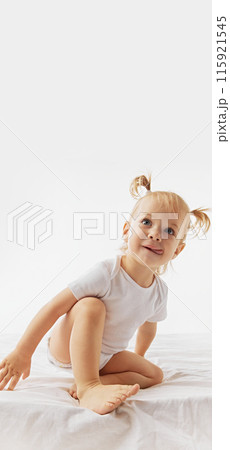 Poster. Blonde toddler with pigtails, wearing onesie sitting on bed against white studio background with negative space to insert your text. 115921545