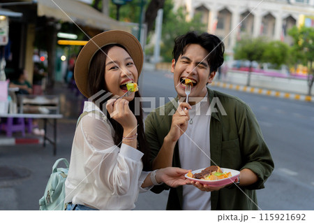Asian couple traveler wearing a casual wear and hat in relationship buying a dessert snack on street food market in the evening. Backpacker traveling in Bangkok, Thailand.. Asian couple traveler wearing a casual wear and hat in relationship buying a dessert snack on street food market in the evening. Backpacker traveling in Bangkok, Thailand.. 115921692
