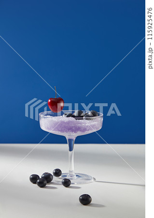 A cocktail glass containing purple grinding ice with some fresh blueberry and a red cherry decorated above, isolated on dark blue background. Blank space for designing and adding text A cocktail glass containing purple grinding ice with some fresh blueberry and a red cherry decorated above, isolated on dark blue background. Blank space for designing and adding text 115925476