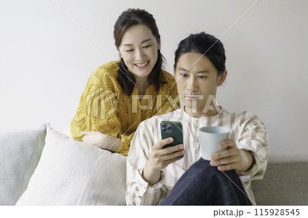 リビングのソファに座ってスマホを見るミドルエイジの男性と後ろからスマホを覗くミドルエイジの女性 115928545