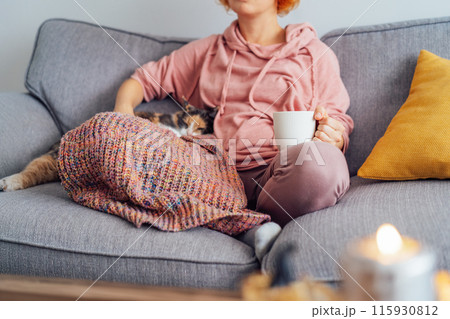 Close up woman in plaid holding cup of tea or coffee, watching movie, TV with multicolored cat on the sofa at home, decorated for fall holidays. Cozy and comfortable autumn concept. Selective focus 115930812