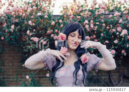 Fairy tale woman portrait. Young beautiful woman Fairy holds pink flower rose on blooming bush background. Hairstyle loose green hair, romantic purple medieval vintage dress. Summer fantasy garden. 115930817