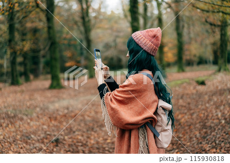Woman wrapped in Shawl Poncho with backpack making photo by phone while enjoying autumn walk in forest. Capture the moment. Feeling harmony. Relaxing, personal fulfillment. Local travel lifestyle. 115930818