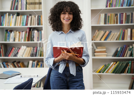 Curly-haired woman standing in the library with a book in hands 115933474