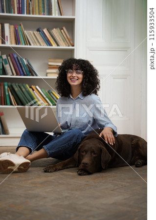 Cute woman working on laptop and stroking her dog lying next to her Cute woman working on laptop and stroking her dog lying next to her 115933504