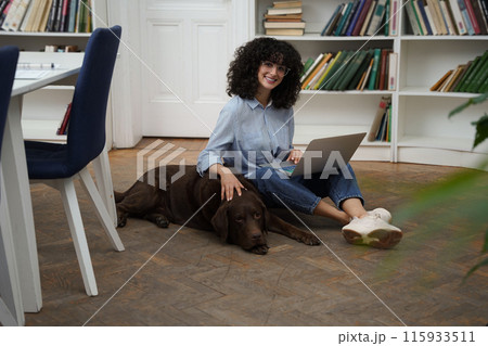 Curly-haired woman with laptop sitting on the floor, a dog next to her 115933511