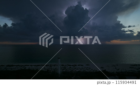 White tall lighthouse on sea shore with blinking light at stormy night for commercial vessels navigation. Thunderstorm with lightnings over ocean water posing danger for ships 115934491