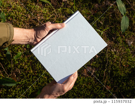 . Hand holding white blank book cover mockup in nature. 115937032