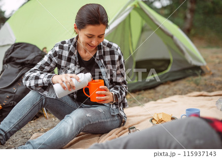 Beautiful adult girl drinking tea in camping in forest with tent on background and smiling. Young woman traveller during hiking in mountains 115937143