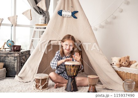 Little drummer girl playing on djembe sitting in front of ethnic tent at home 115937490
