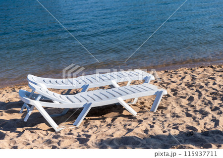 Two white plastic sun loungers stand on a sandy beach Two white plastic sun loungers stand on a sandy beach 115937711
