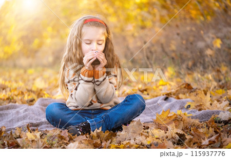 Nice little girl with cup sitting on picnic mat in autumn park Nice little girl with cup sitting on picnic mat in autumn park 115937776
