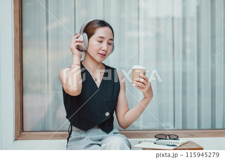 A young woman wearing headphones, holding a coffee cup, and relaxing by the window. She is dressed in casual modern fashion, enjoying a peaceful moment. A young woman wearing headphones, holding a coffee cup, and relaxing by the window. She is dressed in casual modern fashion, enjoying a peaceful moment. 115945279