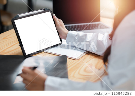 Business professional using a digital tablet at a desk with a laptop and documents in a modern office setting. 115945288