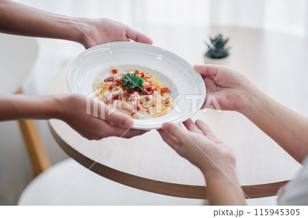 Close-up of hands passing a plate of pasta with bacon and parsley in a bright, minimalist dining setting. 115945305