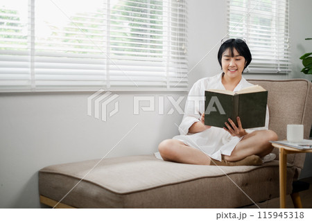 A woman enjoys a peaceful moment reading a book on a cozy sofa in a well-lit, modern living room with large windows and white blinds. A woman enjoys a peaceful moment reading a book on a cozy sofa in a well-lit, modern living room with large windows and white blinds. 115945318