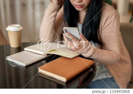A cropped image of an Asian woman using her smartphone at a table indoor while reading books. 115945808
