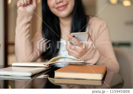 A cropped image of an Asian woman using her smartphone at a table indoor while reading books. 115945809