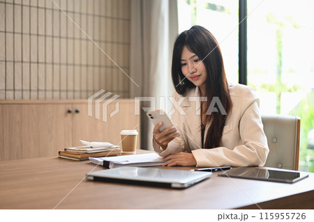 Asian businesswoman sitting at modern workplace with documents and using mobile phone 115955726