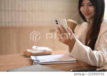An attractive businesswoman drinking coffee and using smartphone at desk, checking social media or communicating online 115955733