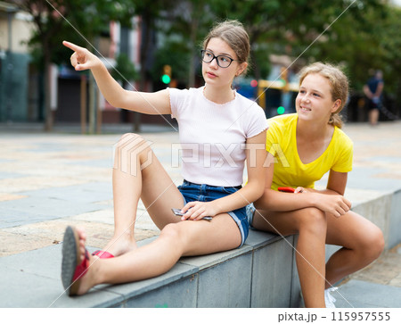 Two girlfriends are sitting on street parapet. One of them points with her hand to something 115957555