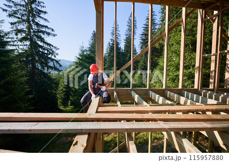 Carpenter constructing wooden frame, two-story house near forest. Man hammering nails with hammer while wearing protective helmet and work coveralls. Concept of eco-friendly modern construction. 115957880