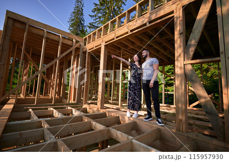 Man and woman examining their future wooden frame dwelling nestled near the forest. Youthful couple at construction site in early morning. Concept of contemporary ecological construction. 115957930