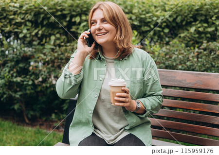 Happy redhead woman in fashion clothes talking on Phone with eco cup of tea. Mockup. Beautiful stylish girl calling on smartphone and sitting on the bench outdoor, hold coffee cup take away. Happy redhead woman in fashion clothes talking on Phone with eco cup of tea. Mockup. Beautiful stylish girl calling on smartphone and sitting on the bench outdoor, hold coffee cup take away. 115959176
