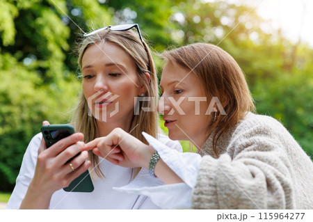 Two young adult happy smiling woman friends portrait using mobile phone smartphone outdoors. Female person people blogger girlfriend watching video streaming chating social media cell at city park Two young adult happy smiling woman friends portrait using mobile phone smartphone outdoors. Female person people blogger girlfriend watching video streaming chating social media cell at city park 115964277