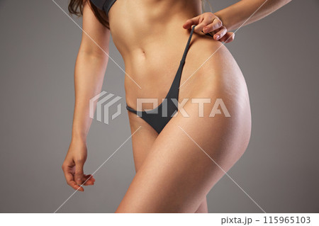 Close-up of woman's lower torso in black bikiny, showcasing her slim and toned body against warm grey studio background. Close-up of woman's lower torso in black bikiny, showcasing her slim and toned body against warm grey studio background. 115965103