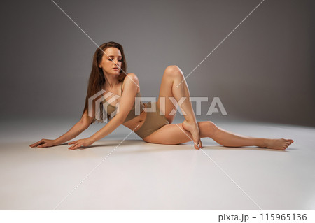 Emphasizing body care, woman's glowing skin and serene expression in beige underwear reflect her wellness regimen against warm grey studio background. 115965136