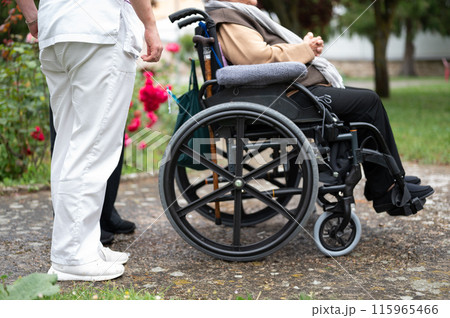 Senior woman in wheel chair with relative and nurse caregiver, outdoors at nursing home garden. 115965466