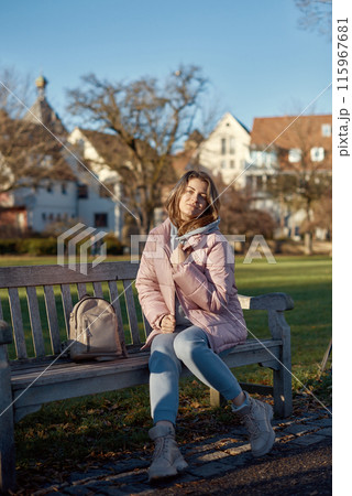Winter Joy in Bitigheim-Bissingen: Beautiful Girl in Pink Jacket Sitting Amidst Half-Timbered Charm. beautiful girl in a pink winter jacket sitting on a bench in a park, set against the backdrop of 115967681