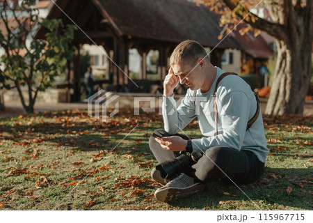 Professional Photographer Taking Picture Of Beautiful Autumn Park. Man Professional Photographer Sit With Camera And With Smartphone In Autumn Park. Retouched, Vibrant Colors, Brownish Tones. 115967715