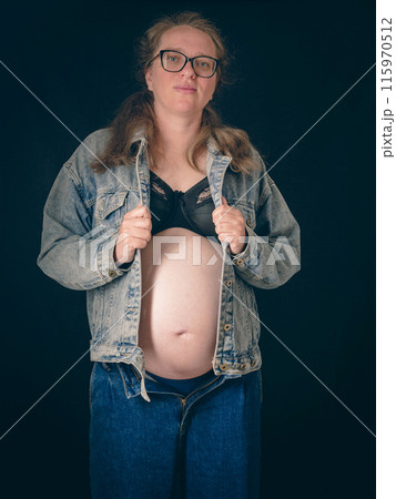An expectant mother stands with her denim jacket open, revealing her pregnant belly. She wears a black top and blue jeans, embodying the anticipation and beauty of motherhood. 115970512